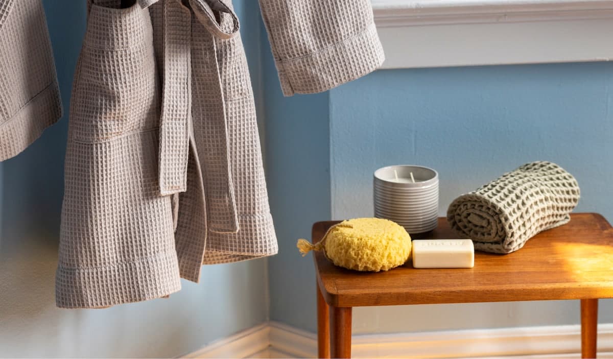 Waffle-weave robes hanging by a blue wall next to a wooden bench with candle, sponge, and soap